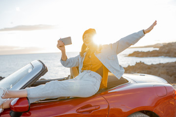 Young stylish woman photographing or vlogging on phone while traveling by car on the rocky coast...
