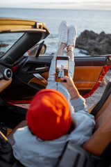 Woman enjoying beautiful sunset view on the ocean, pulling legs out of the car. Carefree travel and...