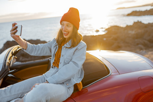 Young stylish woman photographing or vlogging on phone while traveling by car on the rocky coast near the ocean. Lifestyle travel and social influencing concept