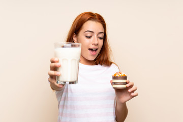 Teenager redhead girl holding a glass of milk and a muffin over isolated background
