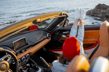 Woman enjoying beautiful sunset view on the ocean, pulling legs out of the car. Carefree travel and...