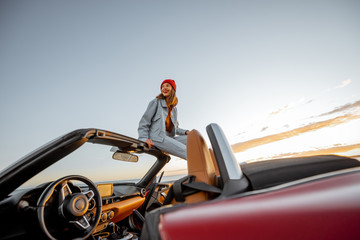 Woman enjoying beautiful view on the ocean, sitting on the car roof top during a sunset. Nature enjoyment and carefree travel by car concept