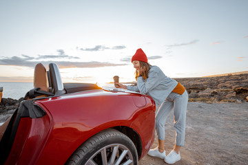 Woman traveling by car near the ocean, leaning against a car trunk, searching some place with a paper map. Beautiful sunset landscape on the background