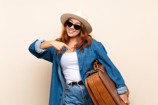 Redhead traveler girl with suitcase over isolated background and pointing it