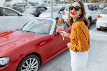 Portrait of a beautiful young woman standing with keys near the red cabriolet at the car parking outdoors. Concept of a happy car buying or renting