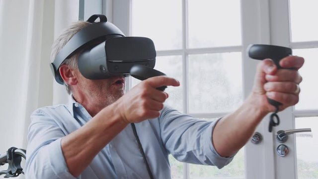 Senior Disabled Man In Wheelchair At Home Wearing Virtual Reality Headset Holding Gaming Controllers