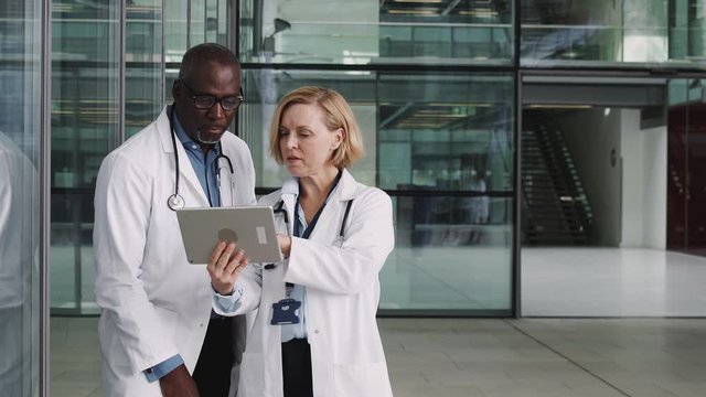 Male And Female Doctors Having Informal Meeting In Modern Hospital Looking At Digital Tablet