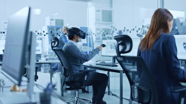 Modern Factory: Industrial Design Engineer Wearing Virtual Reality Headset and Holding Controllers, Uses VR technology for Industrial Design, Development and Prototyping in CAD Software on Computer.