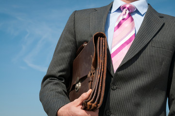 Unrecognizable businessman standing outdoors in gray pinstripe suit with pink tie holding his briefcase in front of a bright blue sky