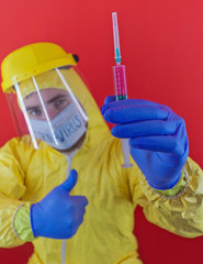 Man in yellow chemical protection suit and face protective mask  with the inscription `coronavirus`. Shoulder portrait. Red background. Without glasses. Protective helmet. Hands in blue gloves. 