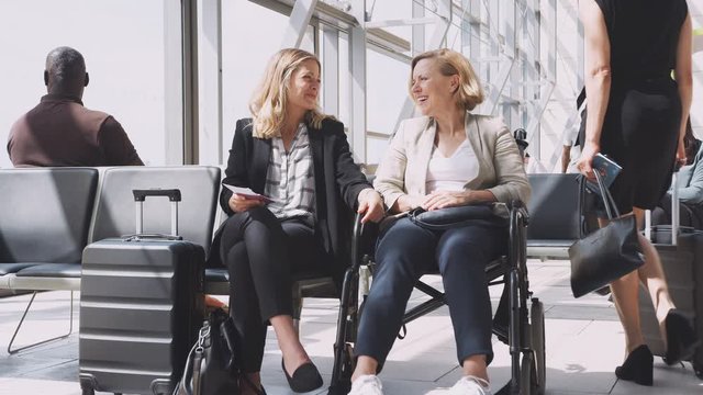 Businesswoman Sitting In Airport Departure With Female Colleague In Wheelchair Talking Together