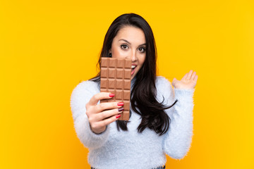 Young Colombian girl over isolated yellow background taking a chocolate tablet and surprised