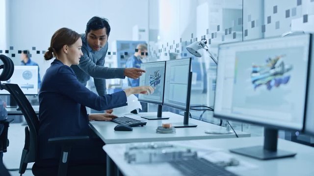 Inside Factory Office: Male Project Supervisor Talks to a Female Industrial Engineer who Works on Computer, Talk. In Workshop: Professional Workers Use High-Tech Industry 4 CNC Machinery, Robot Arm.