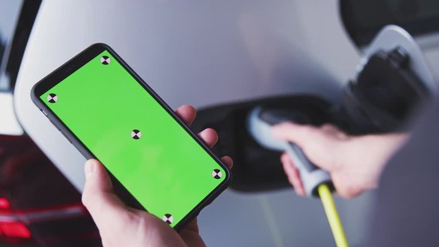 Man Charging Electric Vehicle With Cable Looking At App On Mobile Phone