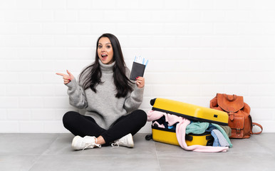 Traveler Colombian girl with a suitcase full of clothes sitting on the floor surprised and pointing finger to the side