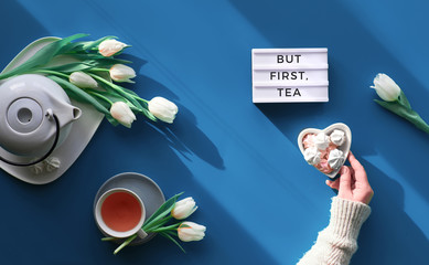 Happy Valentine day, Mother's day or other Spring celebration flat lay. Female hand with heart shape bowl with sweets.. Tea cup, tea pot and white tulips on tale, trendy classic blue background