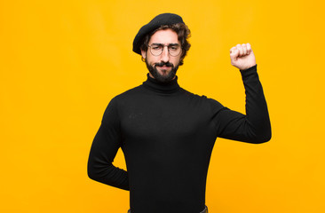 young french artist man feeling serious, strong and rebellious, raising fist up, protesting or fighting for revolution against orange wall