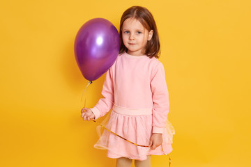 Fototapeta na wymiar Horizontal indoor picture of sweet positive good looking little girl standing isolated over yellow background, holding air baloon in one hand, looking directly at camera, wearing pink dress.