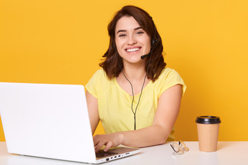 Inddor shot of beautiful young woman wearing t shirt in office in front of white lap top, looking smiling directly at camera, drinhking coffee or tea while working online. Mopdern technology concept.