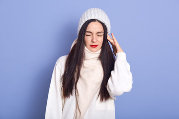 Horizontal picture of young attractive woman standing isolated over blue background in studio, putting hand on ear, having earache, having caught cold, closing eyes, wearing white warm clothes.