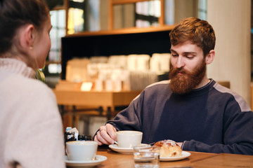Young attractive bearded man drinking coffee while resting with girlfriend in cafe