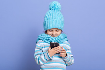 Portrait of attentive adorable little girl wearing blue hat and scarf, striped sweatshirt, standing isolated over lilac background, holding smartphone in both hands, using device. Tech concept