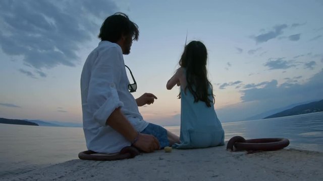 Rear view of little girl sitting on the edge of a dock and fishing with father Mediterranean Sea at sunset. Enjoying adventure in summer vacation	