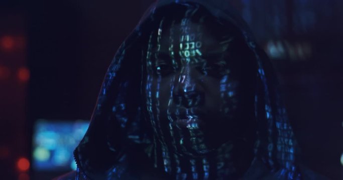 Close up of the young African American guy hacker in a hood looking straight to the camera with projected code numbers and characters on His Face in the dark room full of computers. Portrait.
