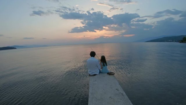 Rear view of little girl sitting on the edge of a dock and fishing with father Mediterranean Sea at sunset. Enjoying adventure in summer vacation	