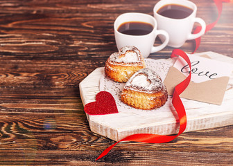 Romantic breakfast for Valentine's Day. Two cups of coffee, two cupcakes in the form of a heart on a white board on a wooden table, a red ribbon, a card with the inscription love. Copy space