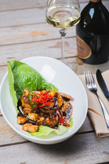 Sliced chicken fried in sweet and sour soy sauce with vegetables and herbs in a deep plate on a wooden table in a restaurant.