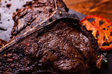 Cooked meat t-bone steak on serving board with garlic cloves, tomatoes, rosemary, spices and glass...