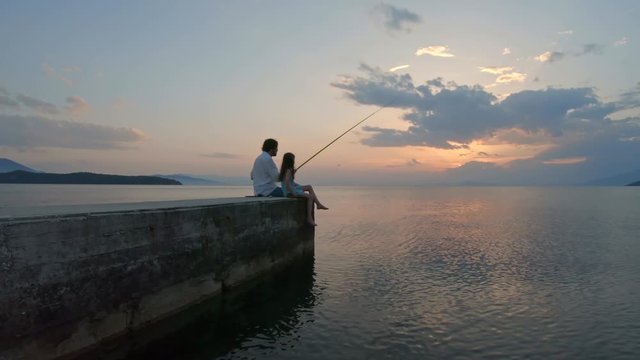 Rear view of little girl sitting on the edge of a dock and fishing with father Mediterranean Sea at sunset. Enjoying adventure in summer vacation	