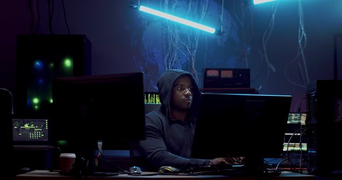 Young African American male online cyber data thief sitting at the computer screen in the dark rom and stealing digital information.