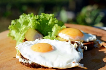 Fried eggs sausage on toast with lettuce on a side dish on a wooden board on nature. Classic summer breakfast.