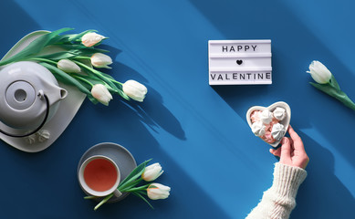 Happy Valentine's day celebration flat lay. Female hands show heart shape sign. Tea cup, tea pot, sweets and white tulips on tale, background in trendy classic blue color.