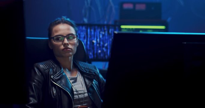Camera zoming in on the Caucasian young software developer woman in glasses sitting in front of the computer screen and resting. At night in the hacker office.