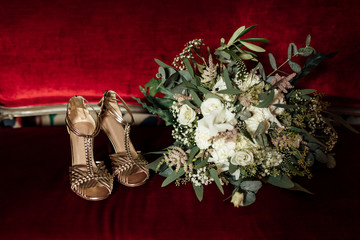 wedding set of bridal bouquet and golden sandals shoes on red classic sofa. bride morning preparation