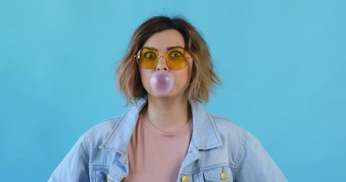 Portrait happy smile woman in yellow sunglasses inflates bubble from chewing gum balloon and bursts it in on blue background. Slow motion. Emotions of people. Lifestyle. Monotone. 