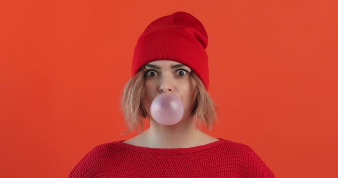 Portrait happy girl in red hat inflates bubble from chewing gum balloon and bursts it in red sweater on red background smiling looking at camera. Monotone. Slow motion. Emotions of people. Lifestyle