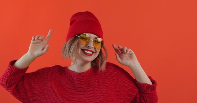Portrait of happy smiling young sexy girl in yellow sunglasses red hat listening to music dancing and waving her hands rhythmically on red background. Pleasure. Emotions people. Monotone. Copy space