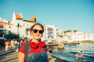 Close up portrait of happy teenage girl posing by the sea, wearing backpack and sunglasses, arms...