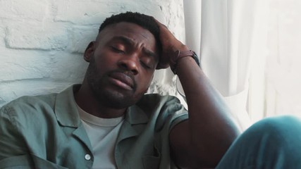 Portrait of sad african american man sitting on windowsill putting hands to the head in home depressed feeling bad annoyed. man tired and having problems close up