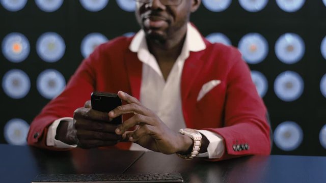 Bearded bald african-american guy dressed in red suit, white shirt and stylish glasses wearing big watch, uses smartphone holding it in hands, in dark office with blurred blue classic background.