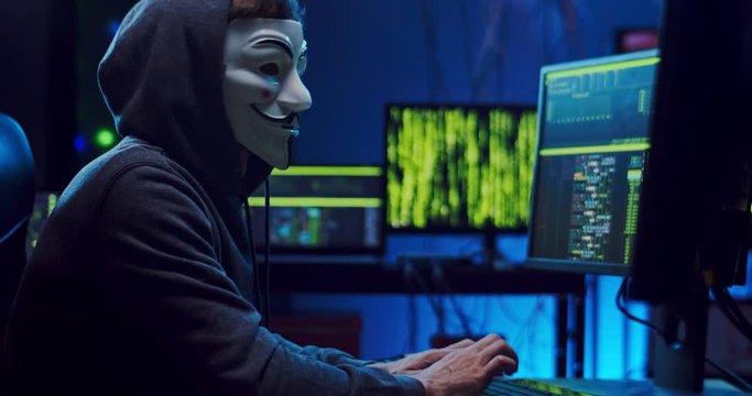Caucasian anonymous hacker man in a mask working and hacking data at the two computers at one time while coding and tapping. At night.