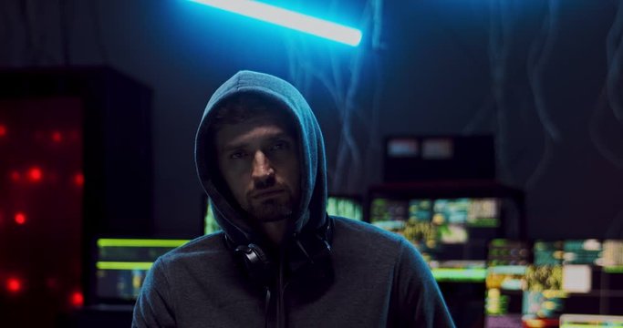 Portrait of the young good looking Caucasian man hacker in a hood looking straight to the camera while sitting in the dark server room with computers. Close up.