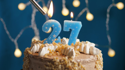 Birthday cake with 27 number candle on blue backgraund set on fire by lighter. Close-up