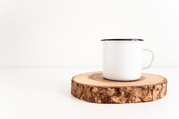 Enamel mug mock up with wooden cut tree section on white table. Boho design of tin cup on white background with copy space for image or text