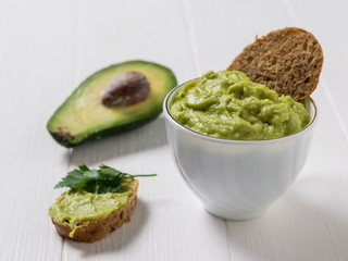Guacamole in a white Cup with thin slices of bread on a white wooden table. Diet vegetarian Mexican food avocado.