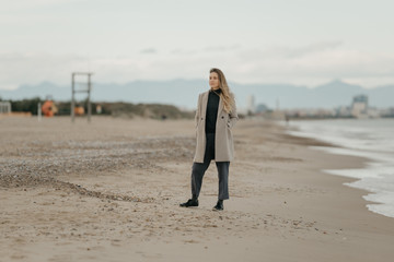A young attractive woman in a coat, trousers, and sneakers in the seashore.